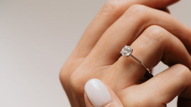 Close up of a solitaire moissanite ring with a slim band, worn on models hand held up against a beige background. 