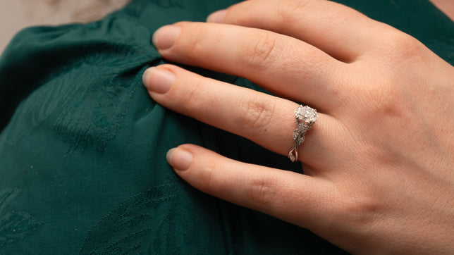 Accented engagement ring in Platinum with oval centre stone and twisted vine design, worn on the hand of model wearing a forest green dress. 