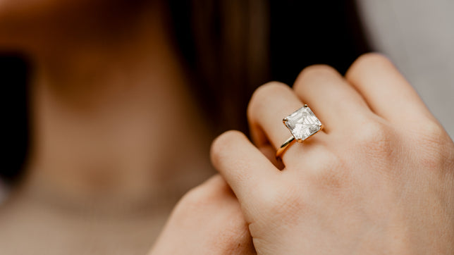 Close up shot of large carat asscher solitaire engagement ring worn on models hand, models neck and long dark hair can be seen out of focus in background. 