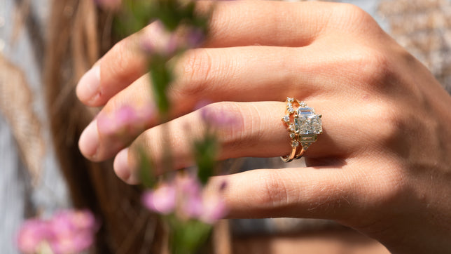 Emerald accented engagement ring stacked with a scattered multi-stone wedding band is worn on woman's hand, photographed outside in sunlight with out of focus pink flowers in foreground. 