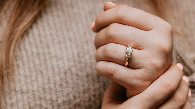 Trilogy gold moissanite engagement ring worn on models hand, model is wearing a grey knit jumper and has light pink painted nails. 