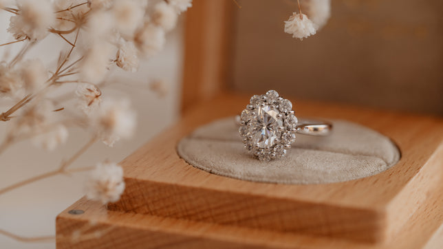 Close up of vintage inspired oval halo engagement ring in Platinum, photographed on an open wooden ring box with dried white flowers to the left hand side. 