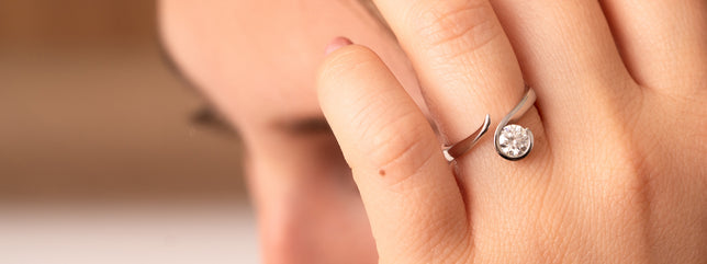 Close up of modern solitaire engagement ring set with a round brilliant lab-grown diamond, worn on woman's hand, her face and hair can be seen in background. 