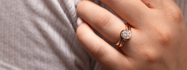 Close up shot of modern style engagement ring with round brilliant centre and an accented band, worn on models hand holding dried flowers. 