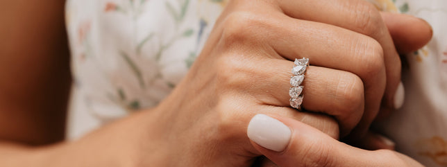 Five stone pear cut moissanite ring worn on models hand, models white flowery dress can be seen in background. 