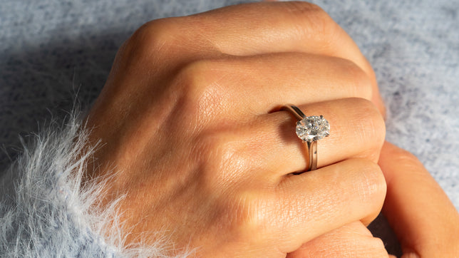 Close up of oval lab diamond solitaire engagement ring with platinum band worn on models hand with models light blue fluffy jumper in background. 