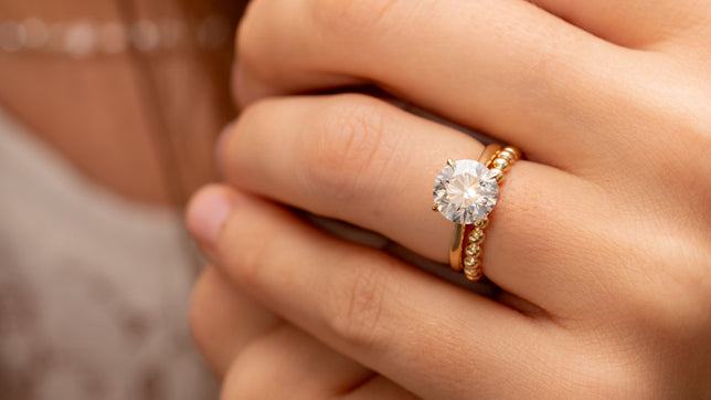 Close up of woman wearing a round brilliant solitaire engagement ring in yellow gold stacked with a yellow gold beaded band. The models white lacy top and brown hair can be seen in background. 