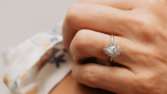 Close up of vintage inspired style pear cut halo engagement ring in Platinum is worn on women's hand. Her hand is resting on her shoulder and her white flowery summer dress can be seen in background. 