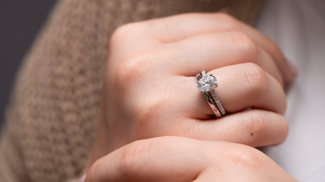 Close up of oval wedding ring friendly shoulder set engagement ring stacked with a plain platinum wedding band, worn on models hand. 